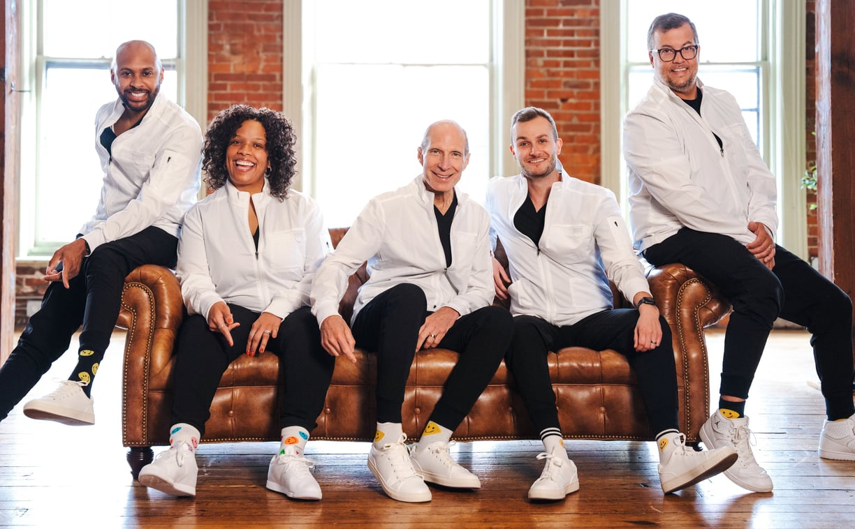 docs-sitting-on-couch-YX4yK52X8yUrgJZe Five people smiling on a leather couch.